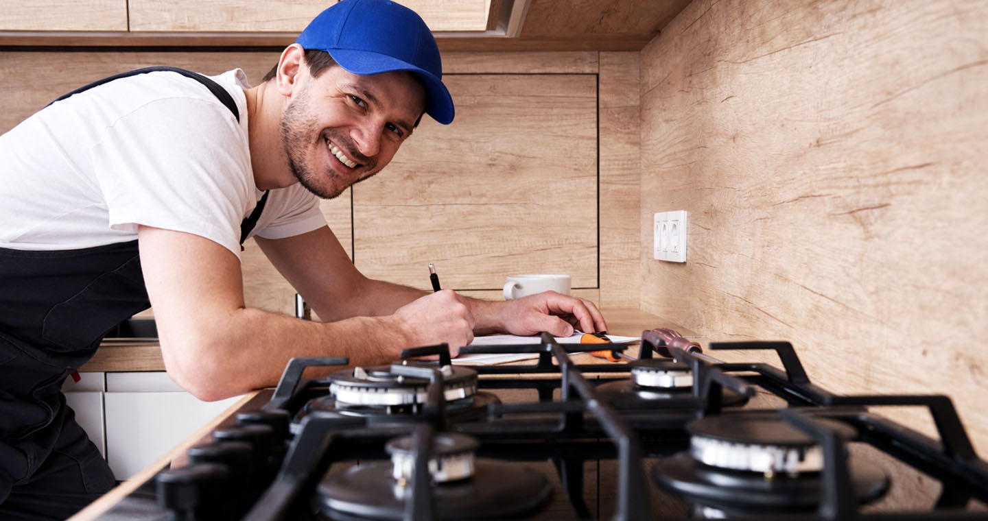 Smiling technician in black overalls and a blue cap writes notes beside a gas cooktop in a modern kitchen
