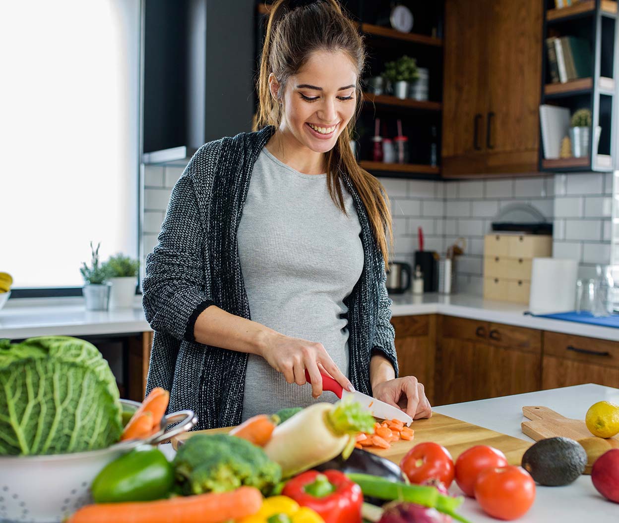 A smiling woman chops carrots on a cutting board in a kitchen surrounded by fresh produce
