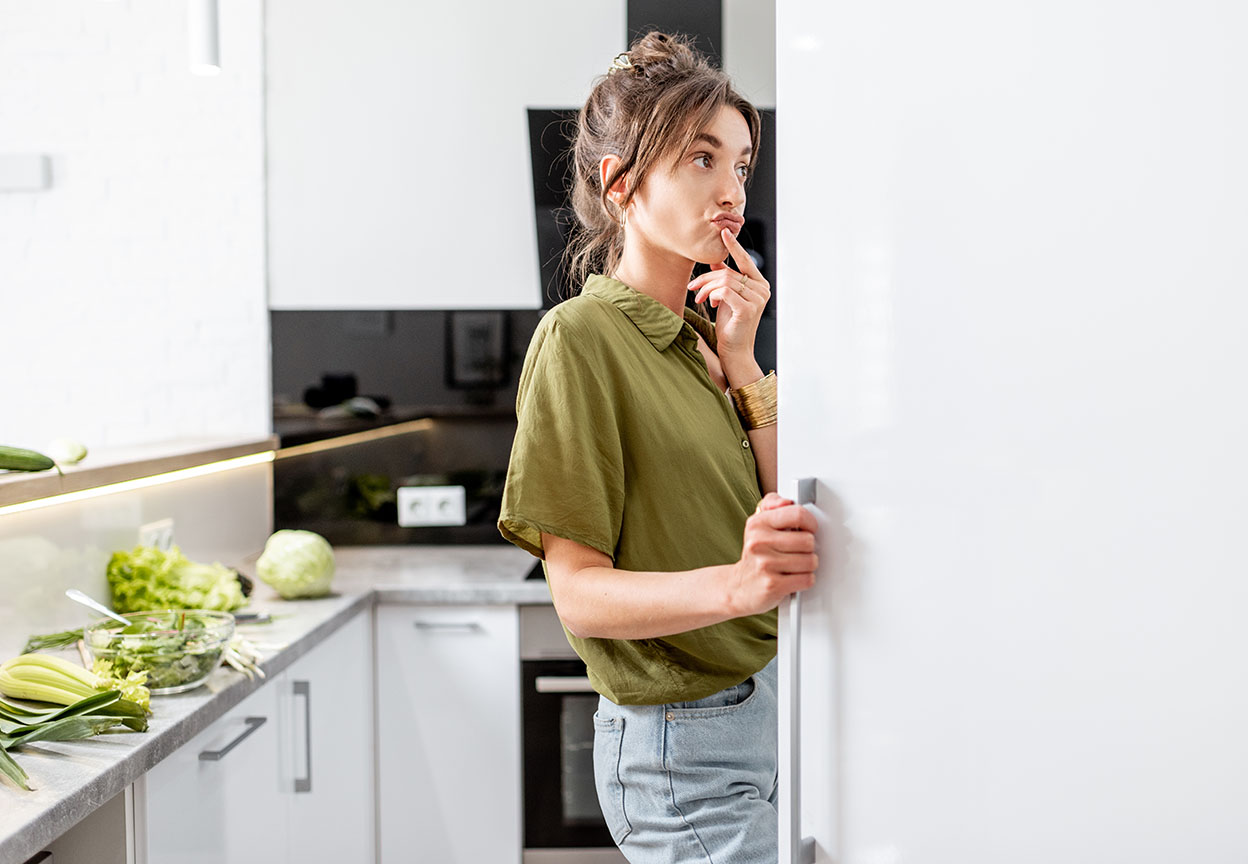 A woman stands at an open refrigerator, thinking while fresh vegetables sit on the kitchen counter