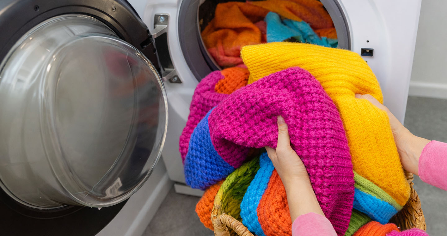 Hands load bright, thick knit sweaters from a basket into an open front-loading washer