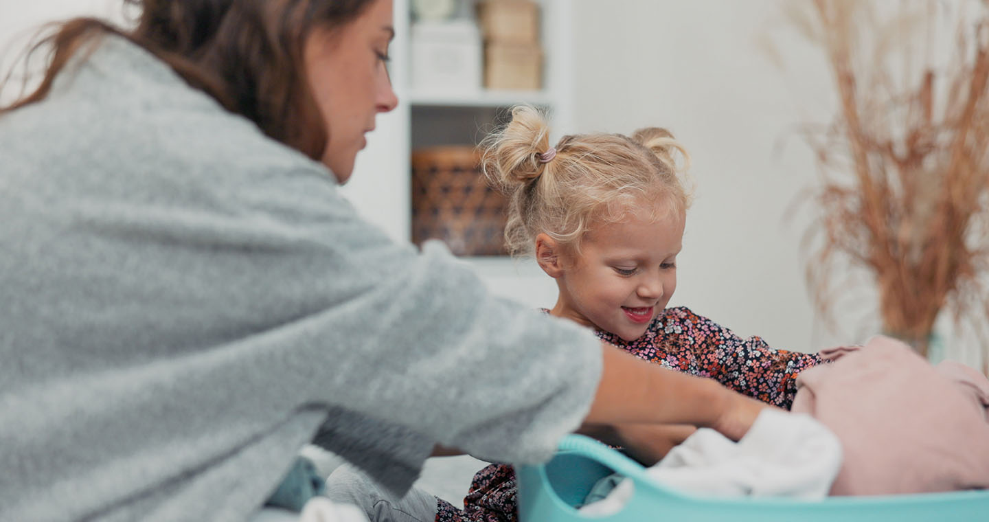 A woman helps a smiling young girl fold clothes into a laundry basket at home