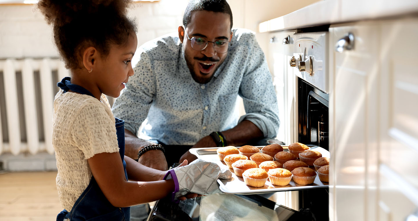 An adult and child pull a tray of freshly baked muffins from the oven in a bright kitchen