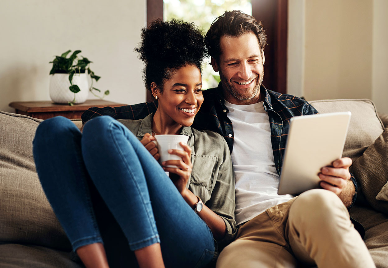 A couple sits on a couch smiling while looking at a tablet and holding a warm drink