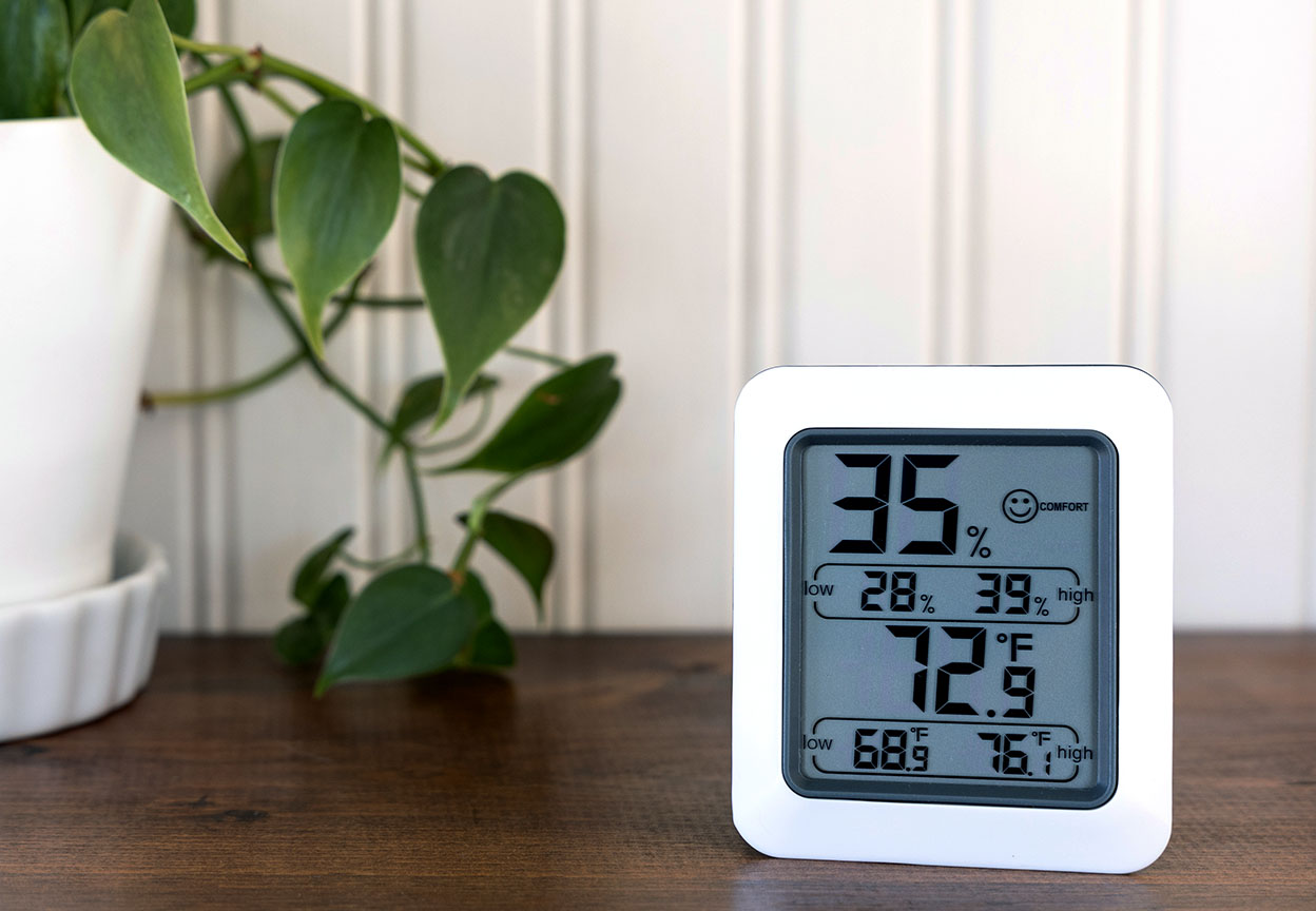 A digital humidity monitor on a table shows the indoor humidity level and temperature beside a plant