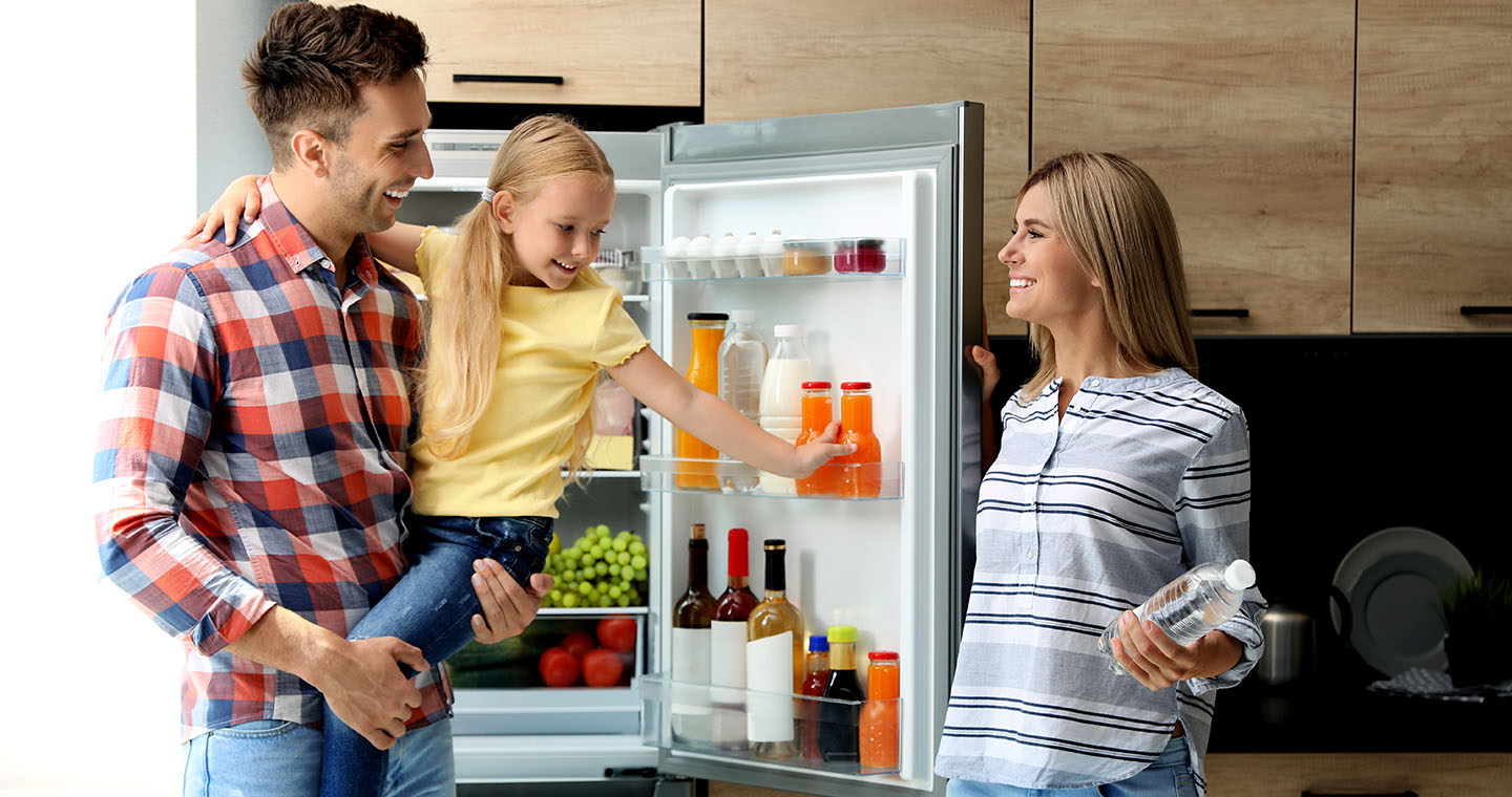 A family smiles in a modern kitchen as a dad holds his daughter while she reaches for a drink in an open refrigerator and mom stands nearby holding a water bottle
