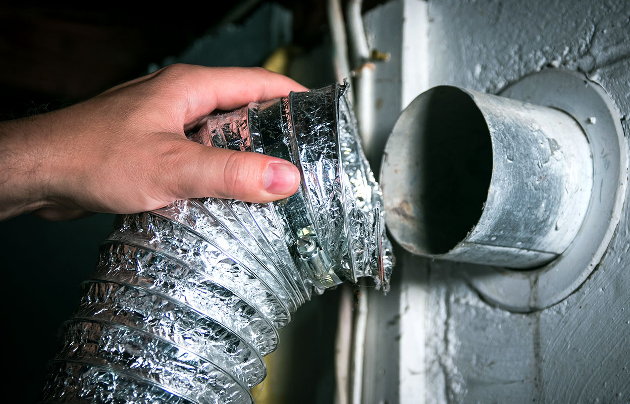 Hand holding a detached flexible dryer vent duct next to a wall exhaust pipe