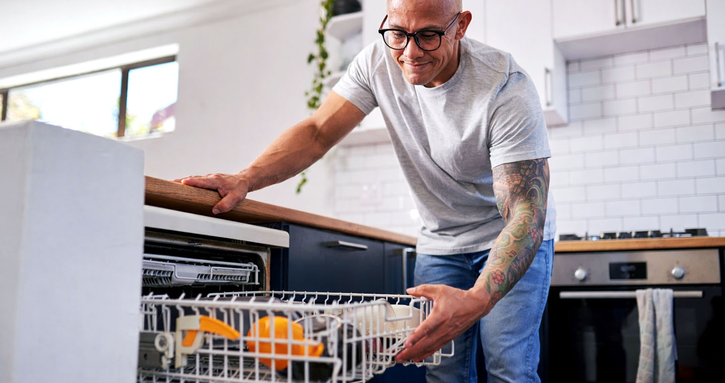 A man with glasses and a tattoo sleeve gently slides out a dishwasher rack in a bright, modern kitchen