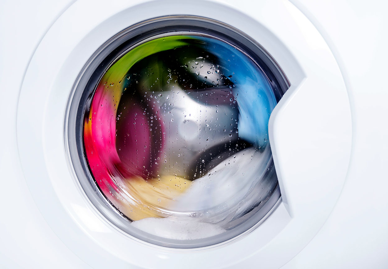 Colorful clothes tumble behind the glass door of a front load washer with water droplets