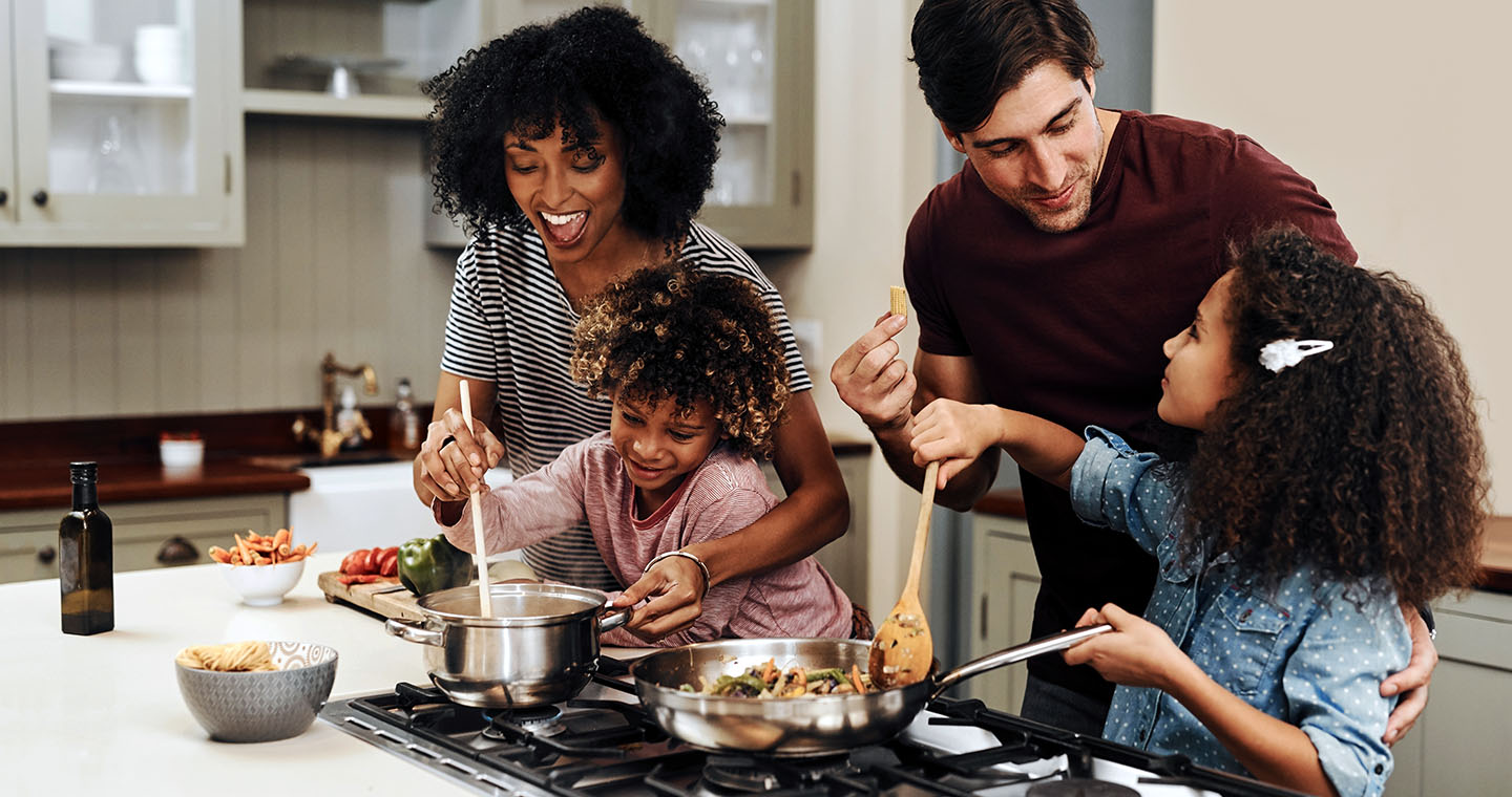 Family cooking together at the stove as kids help stir and taste in the kitchen