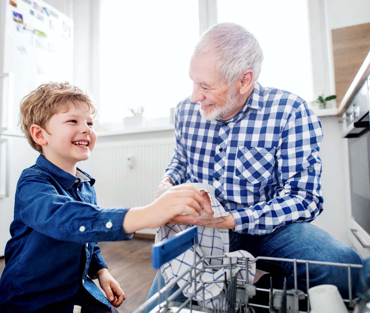 A smiling child hands a dish towel to an older man as they unload a dishwasher together in a bright kitchen