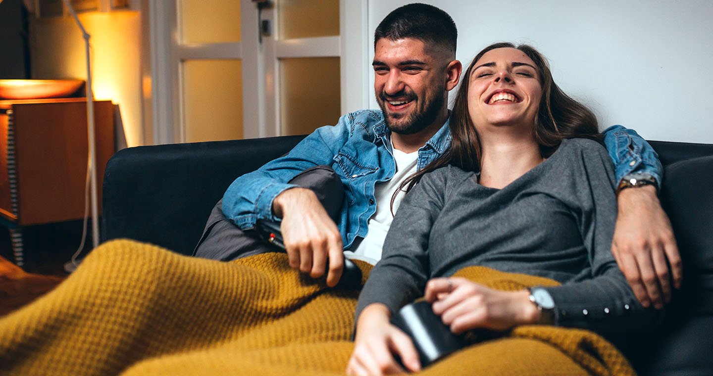 A couple relaxes on a couch under a cozy blanket, laughing together while holding a TV remote.