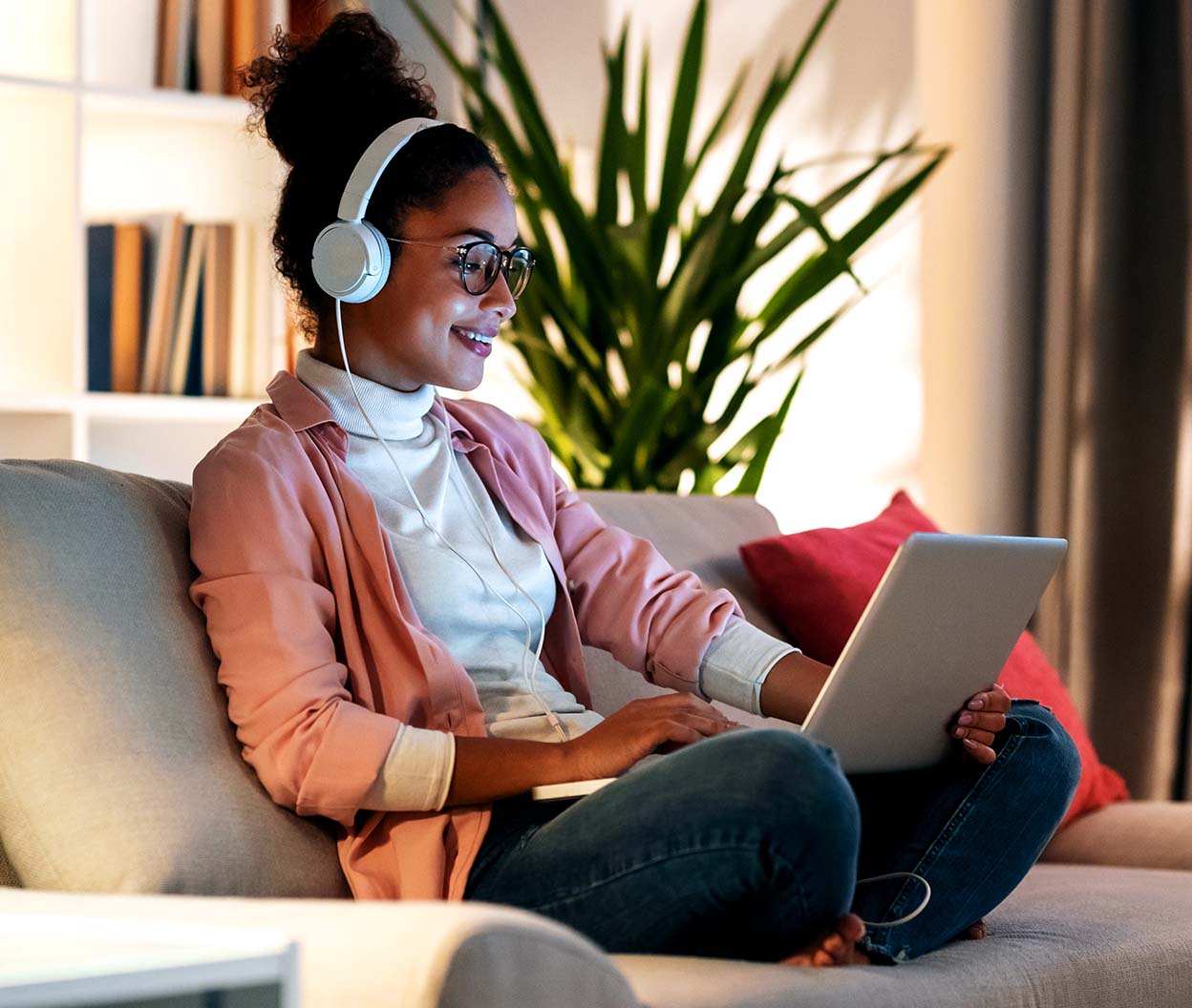 A woman in glasses and headphones sits on a couch using a laptop at home
