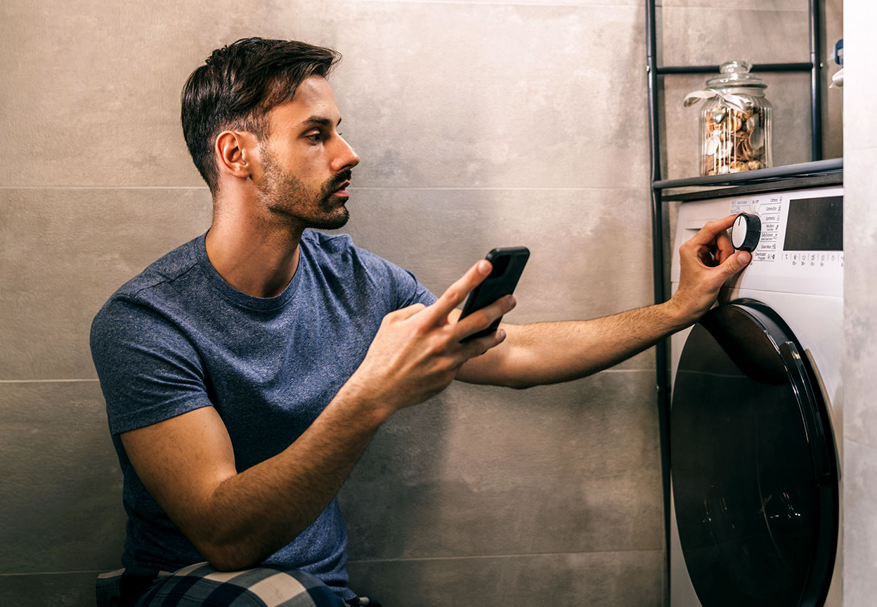 A man checks his phone while turning the dial on a front-loading washing machine