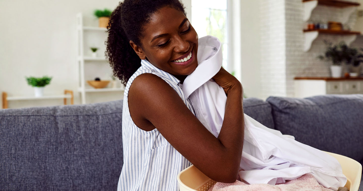 Smiling woman hugs a warm stack of fresh laundry beside a basket on the couch