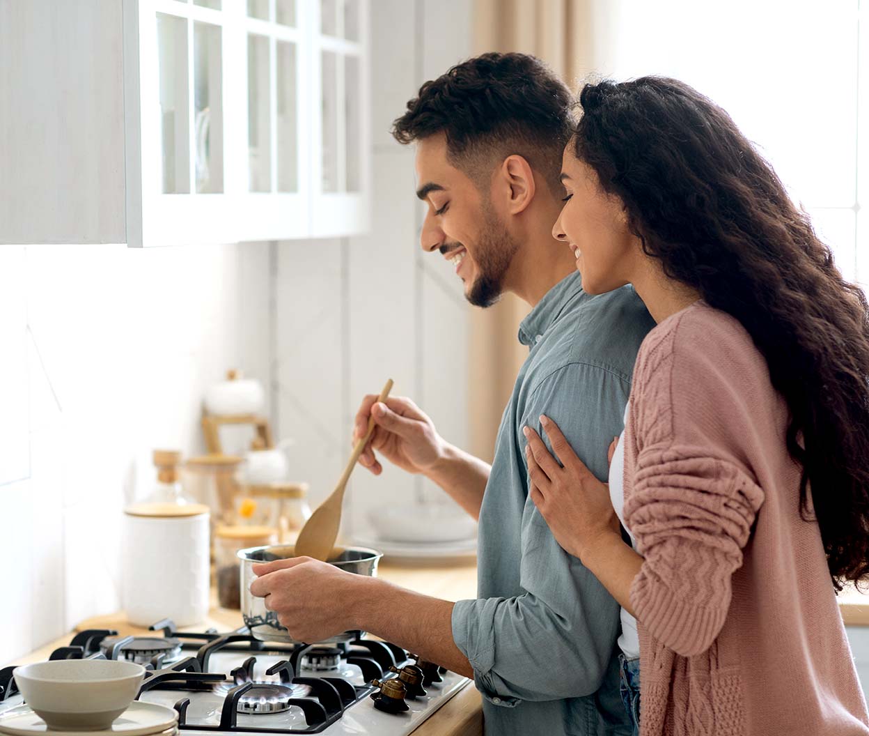 A smiling couple stands at a gas stove in a bright kitchen while cooking together.