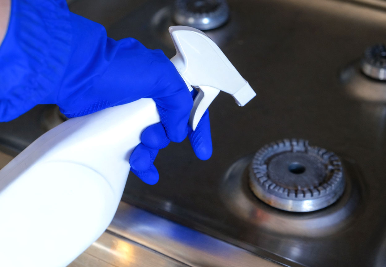 A hand in a blue cleaning glove holds a spray bottle over a stainless steel gas stovetop burner