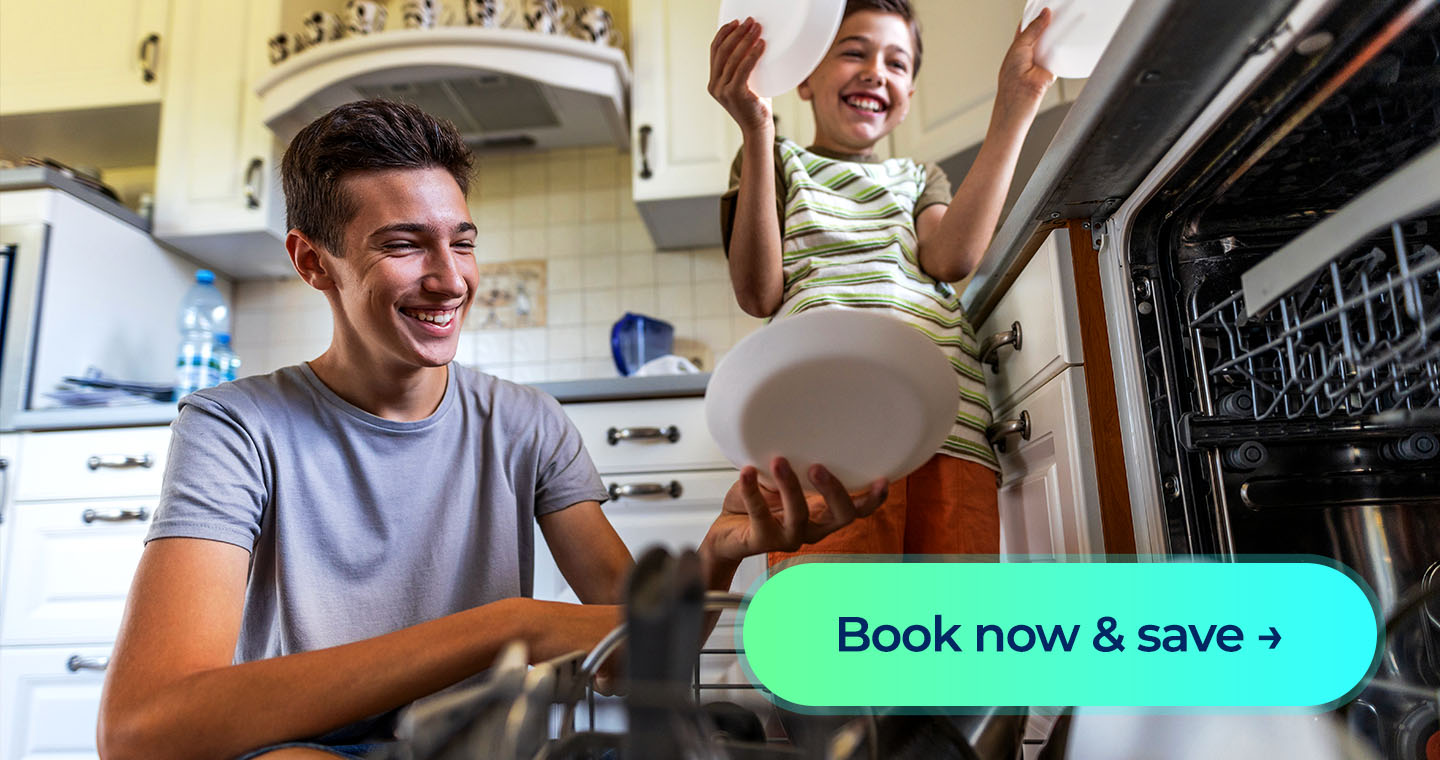 Two kids smile as they load plates into an open dishwasher in a bright kitchen. Button: Book now & save