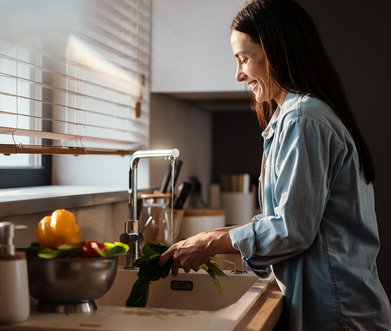 A smiling woman rinses leafy greens at a kitchen sink beside a sunlit window with fresh vegetables on the counter