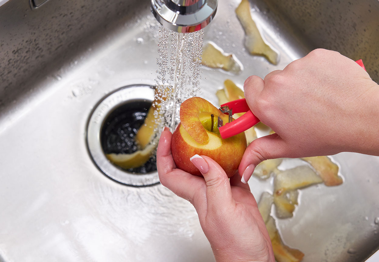 Hands peel an apple under running water above a kitchen sink and garbage disposal with apple peels scattered around the basin