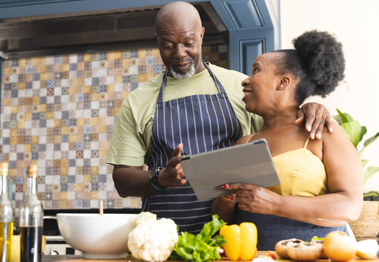 A couple in aprons stands in a kitchen looking at a tablet together with fresh vegetables and cooking ingredients on the counter