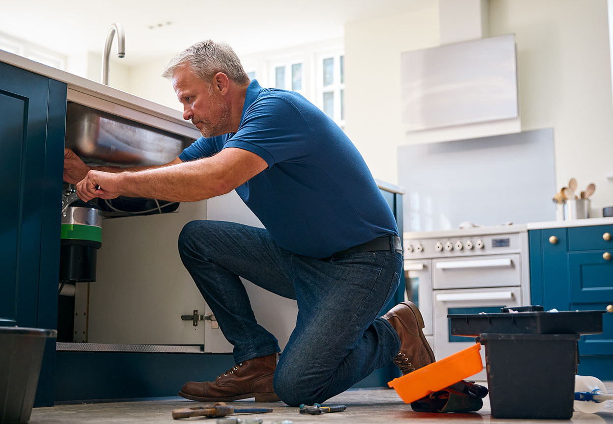 A man kneels on a kitchen floor repairing the plumbing beneath a sink with tools and a bucket nearby