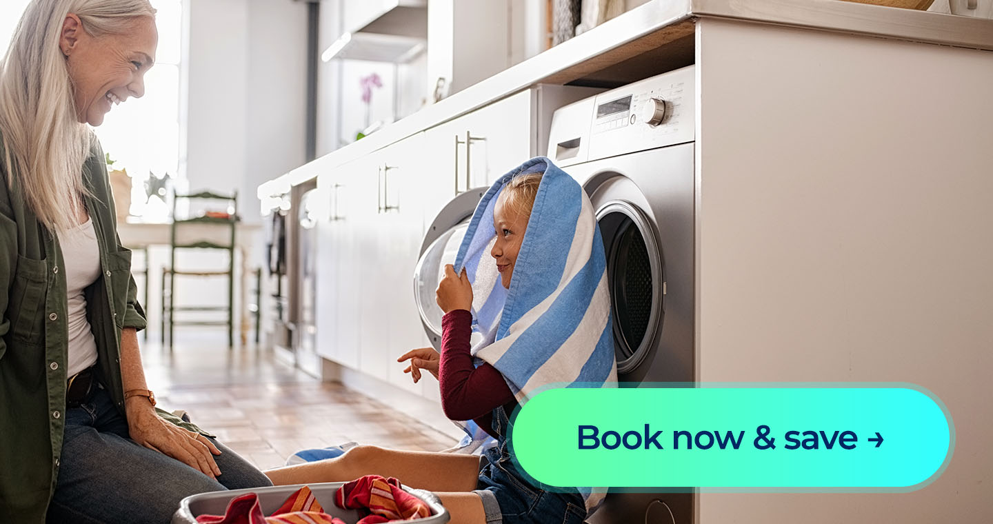 An older woman playing around near a washer with her granddaughter who has a towel over her head