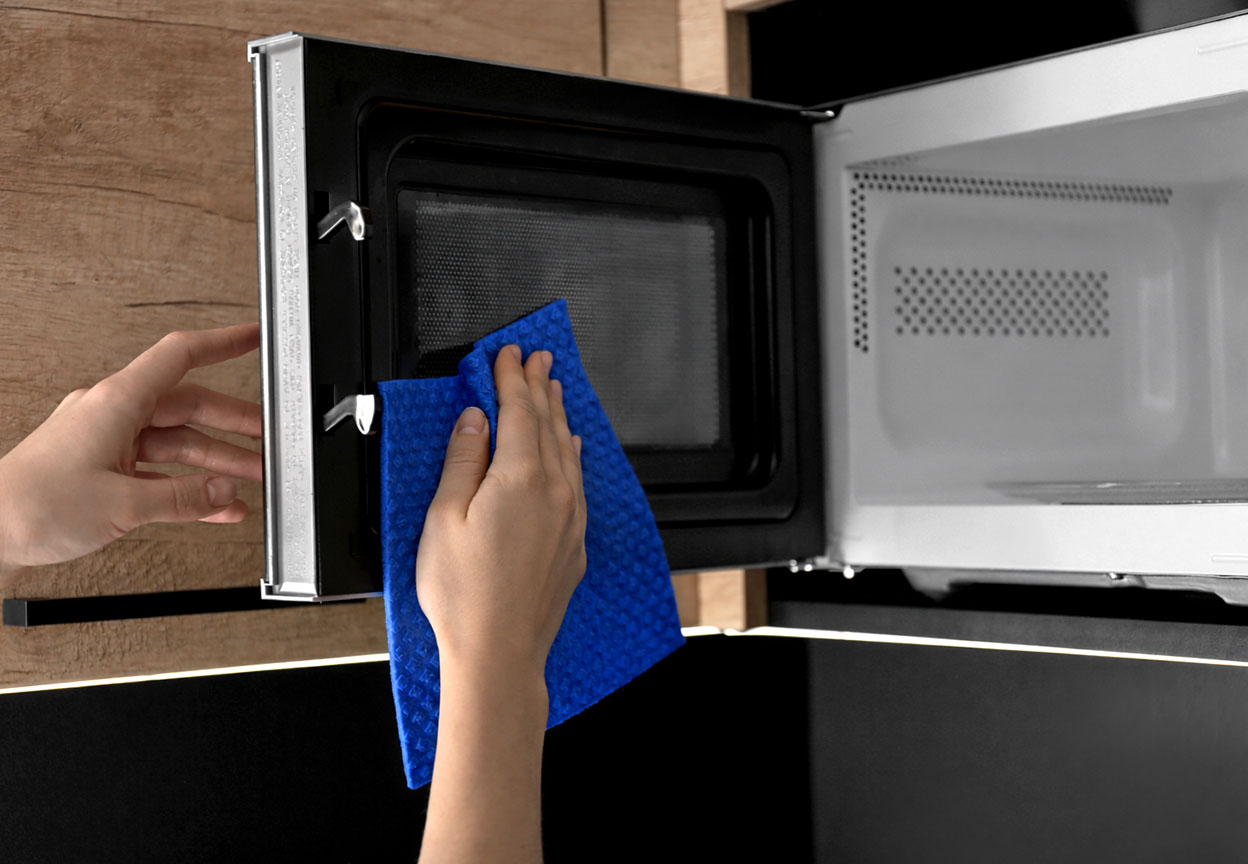 Person wiping the inside of an open microwave door with a blue cleaning cloth