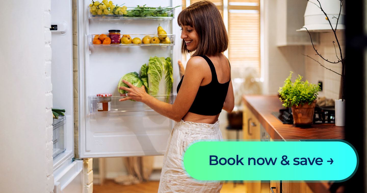 A smiling woman looks into an open fridge filled with fresh produce beside a “Book now & save” button.