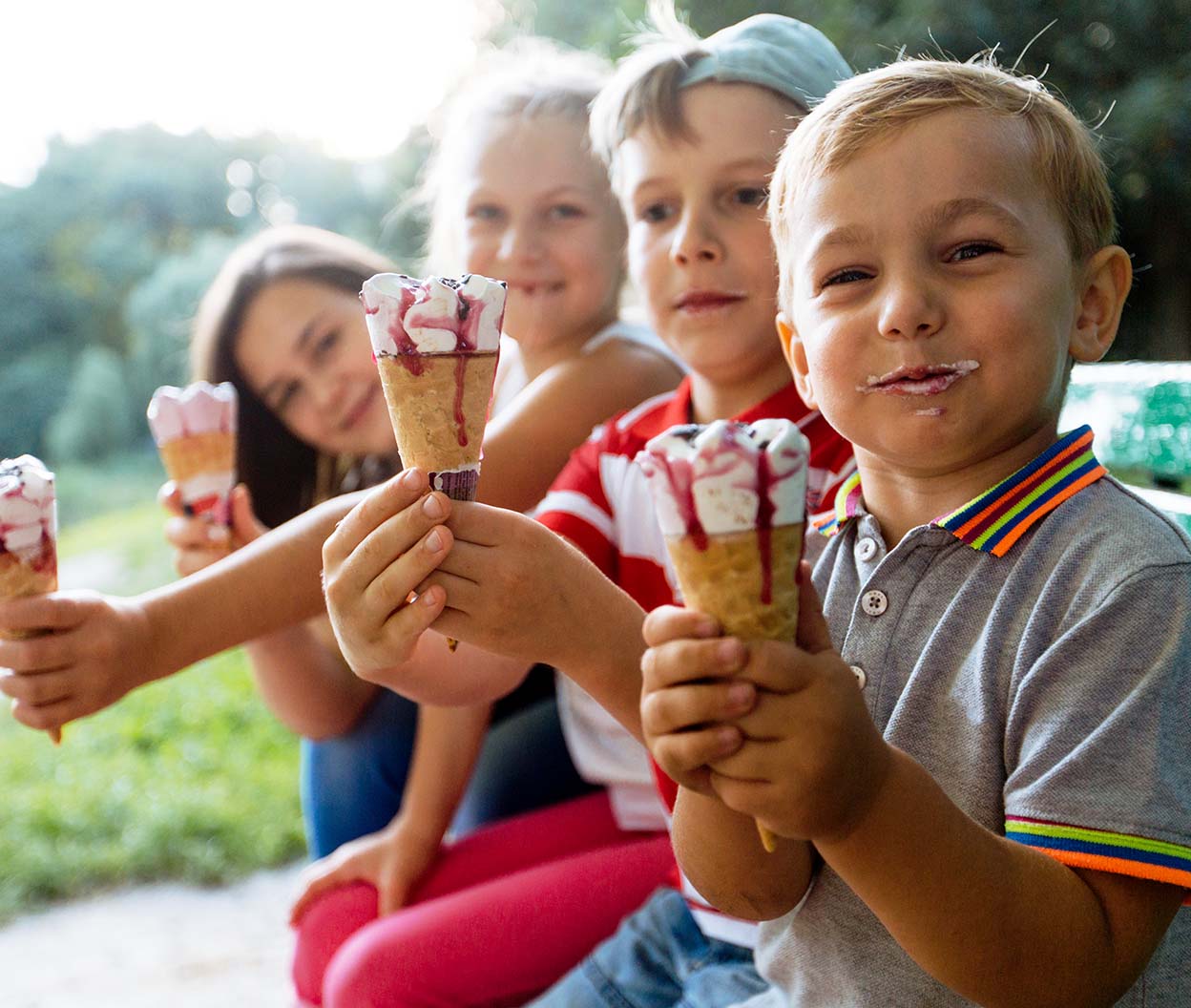 Four children sit together outdoors holding melting ice cream cones and smiling in the summer light