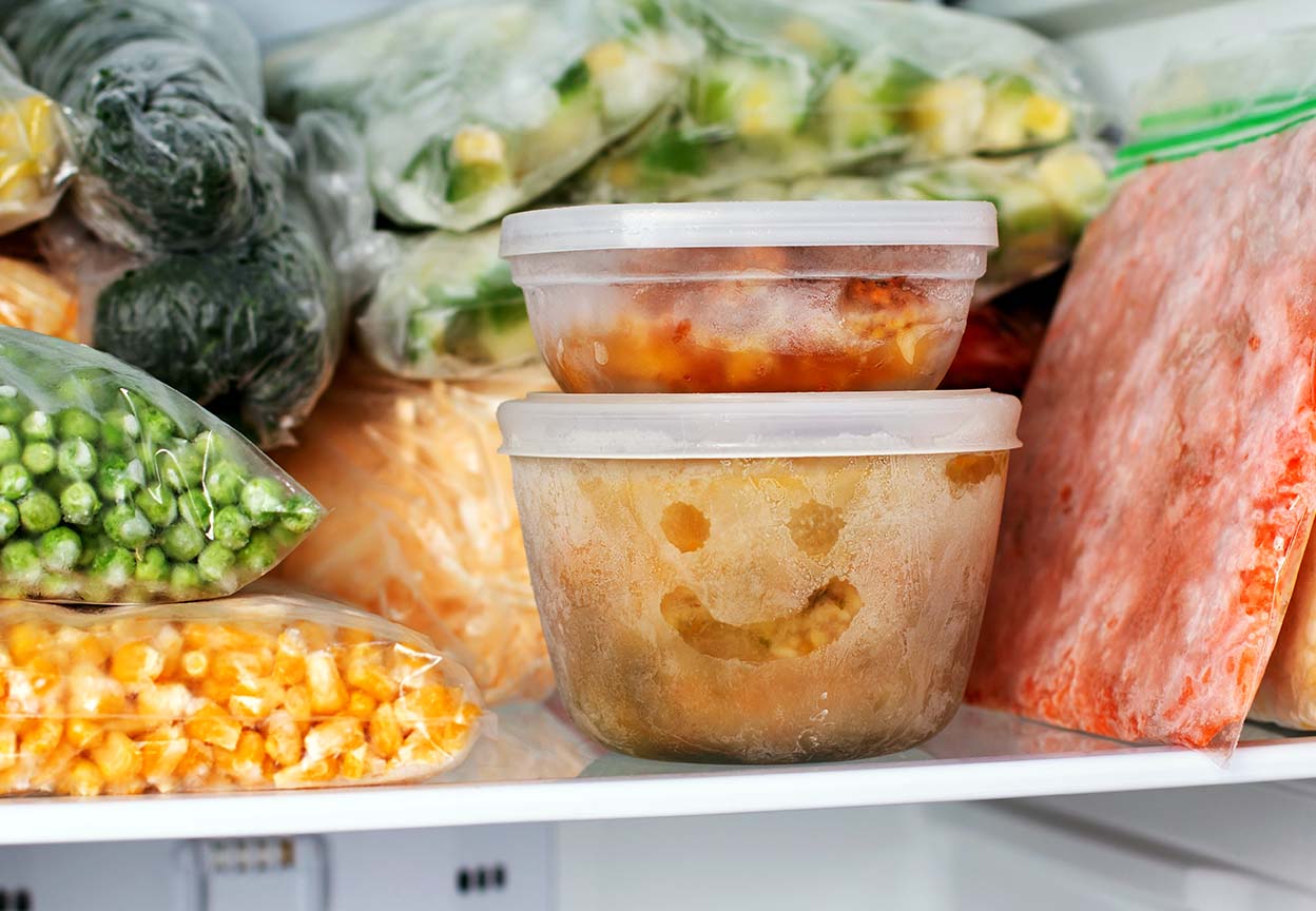 Frozen vegetables and containers fill a freezer, including a frosty food tub with a smiley face on the front