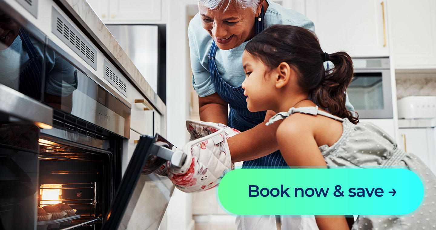 A grandmother pulls a tray of fresh muffins from the oven as her granddaughter watches with excitement beside a “Book now & save” button