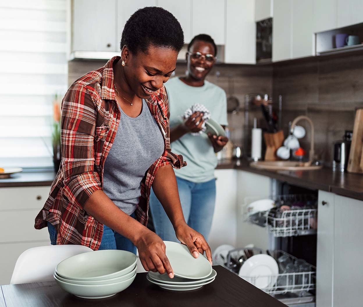 Two people unload clean dishes from an open dishwasher in a bright kitchen
