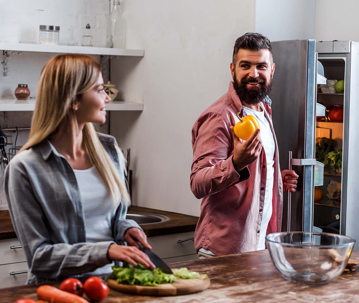 A man holds a yellow bell pepper from an open refrigerator while a woman chops fresh vegetables in the kitchen