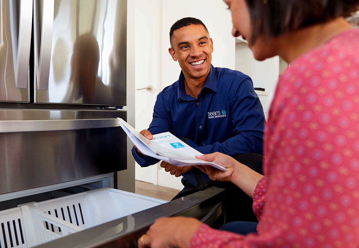 A Sears Home Services technician reviews home warranty paperwork with a homeowner near a refrigerator
