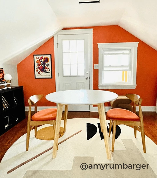 Room with small table with 2 chairs, bookshelf and rug with one wall painted in Emberglow. @amyrumbarger.
