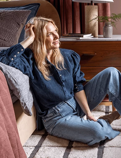 Emily Henderson, looking off to her left, sitting on a carpeted floor and leaning against a sofa with throw pillows & blankets.