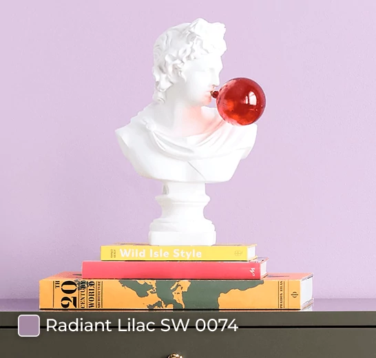 A classical bust sculpture blowing a glass red bubble sitting on top of books against a wall painted in Radiant Lilac SW 0074.