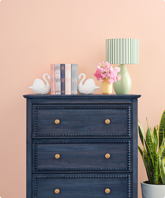 Blue dresser with white swans book ends with books, planter and green lamp in front of peach wall.