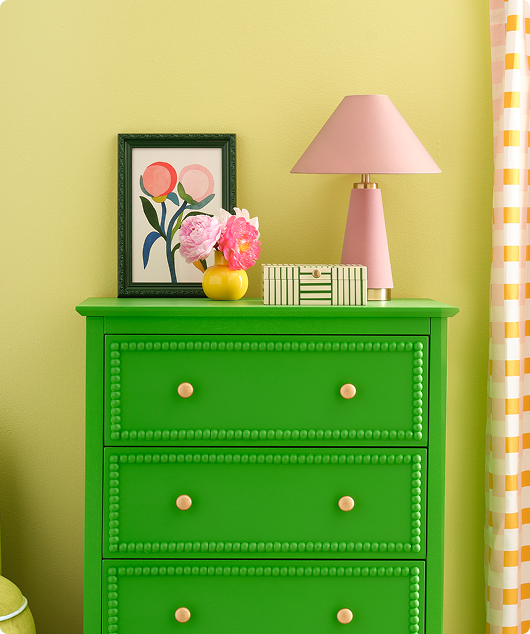 Green dresser, with pink lamp and home decor on top, against yellow wall.