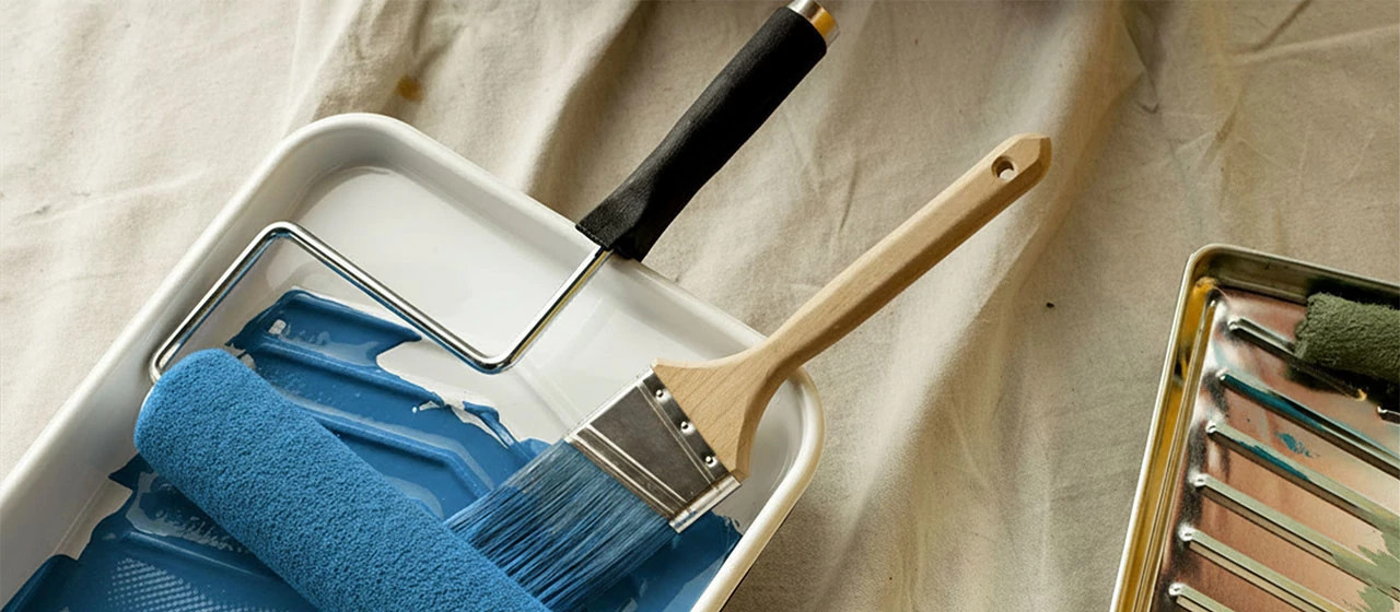 A paint brush and roller on a painting tray with blue paint.