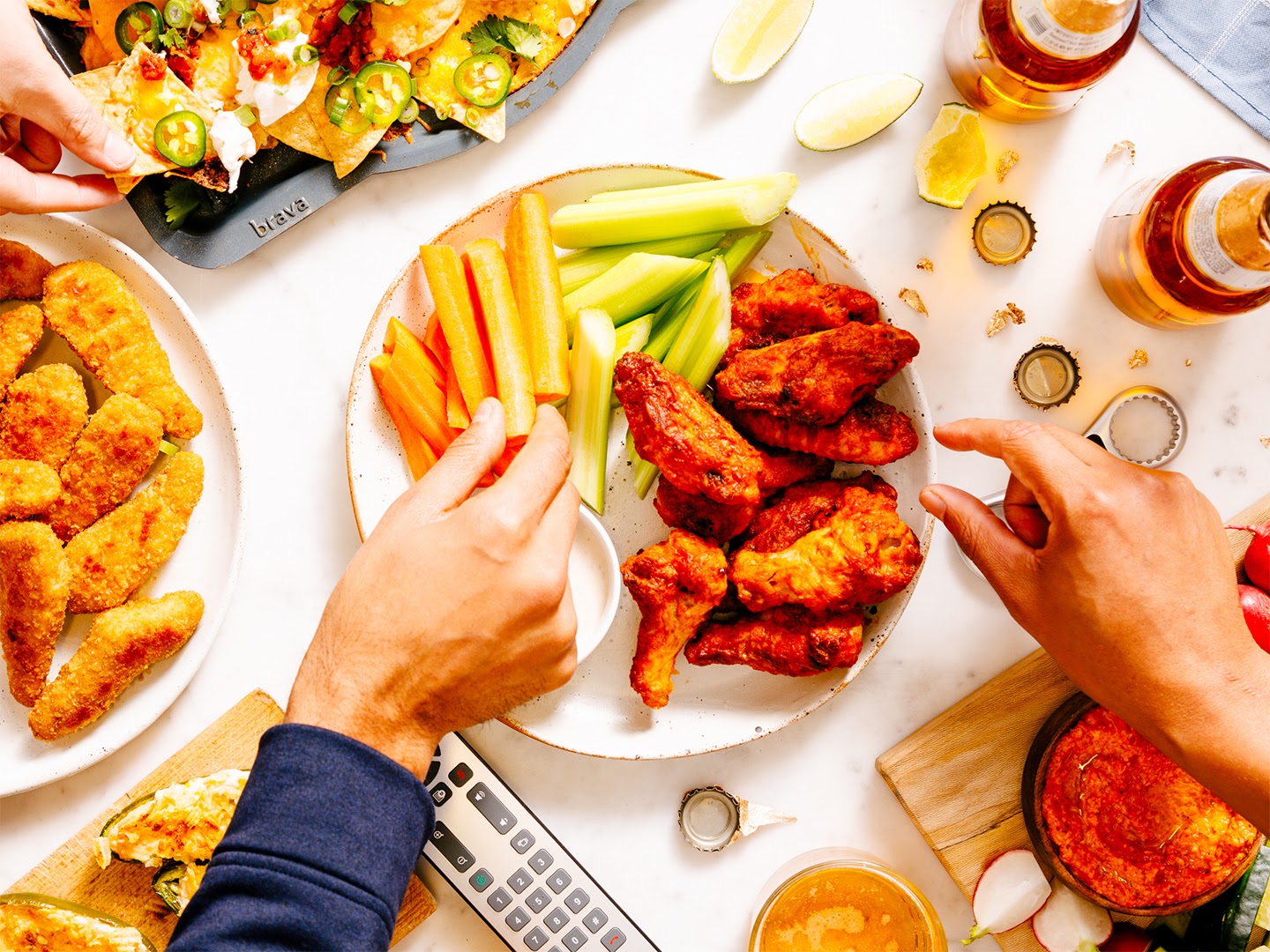 A image of hands picking up pieces of food from a table full of game day recipes like nachos and chicken wings