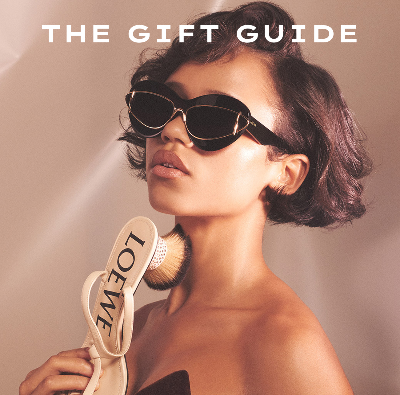 The Gift Guide Hero Image Featuring a Woman Wearing Black Designer Sunglasses and Holding a Loewe Sandal Near Her Neck