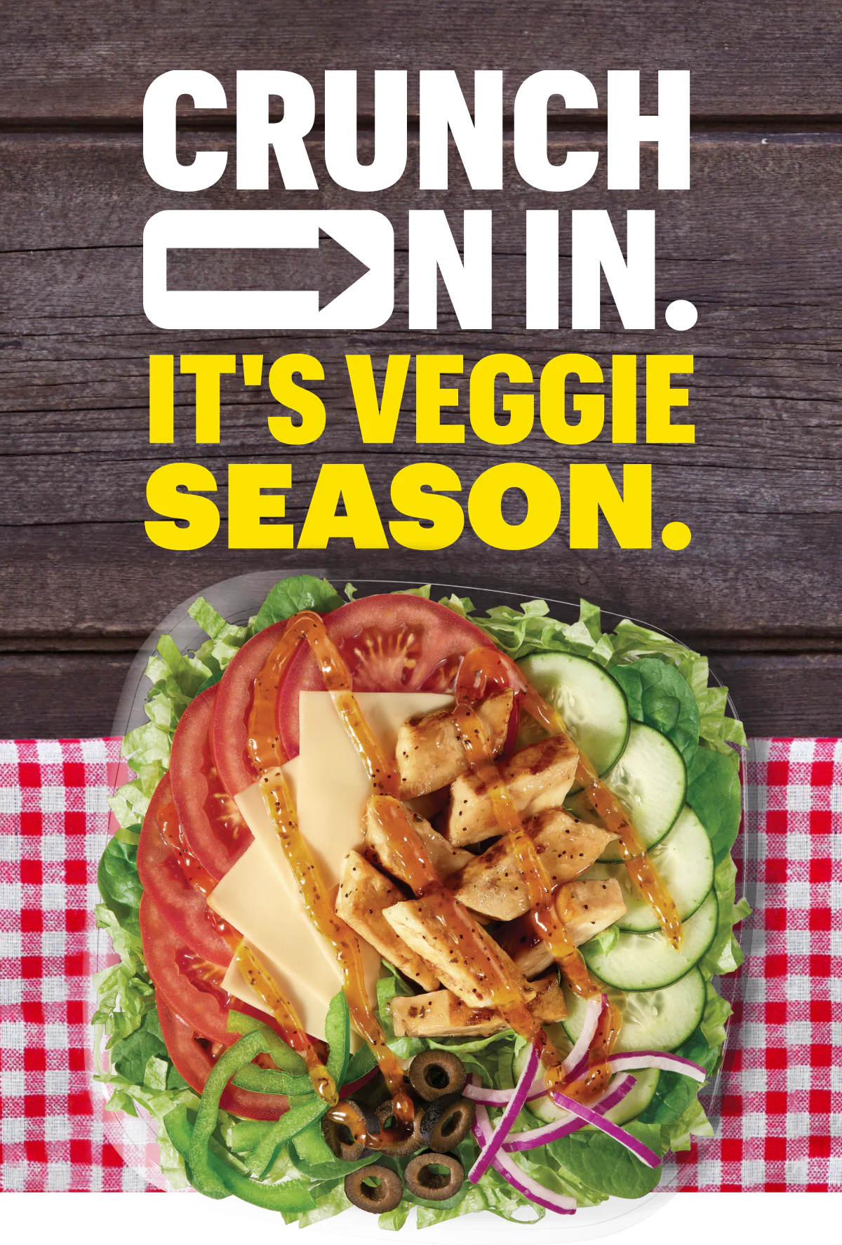 A salad with lettuce, tomato, green pepper, onion, olive, cheese, chicken and dressing sits on a red gingham placemat atop a dark wooden table. On top are the words, "Crunch on in. It's veggie season."