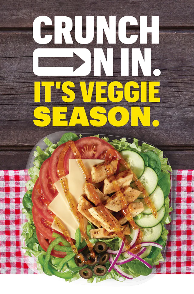 A salad with lettuce, tomato, green pepper, onion, olive, cheese, chicken and dressing sits on a red gingham placemat atop a dark wooden table. On top are the words, "Crunch on in. It's veggie season."