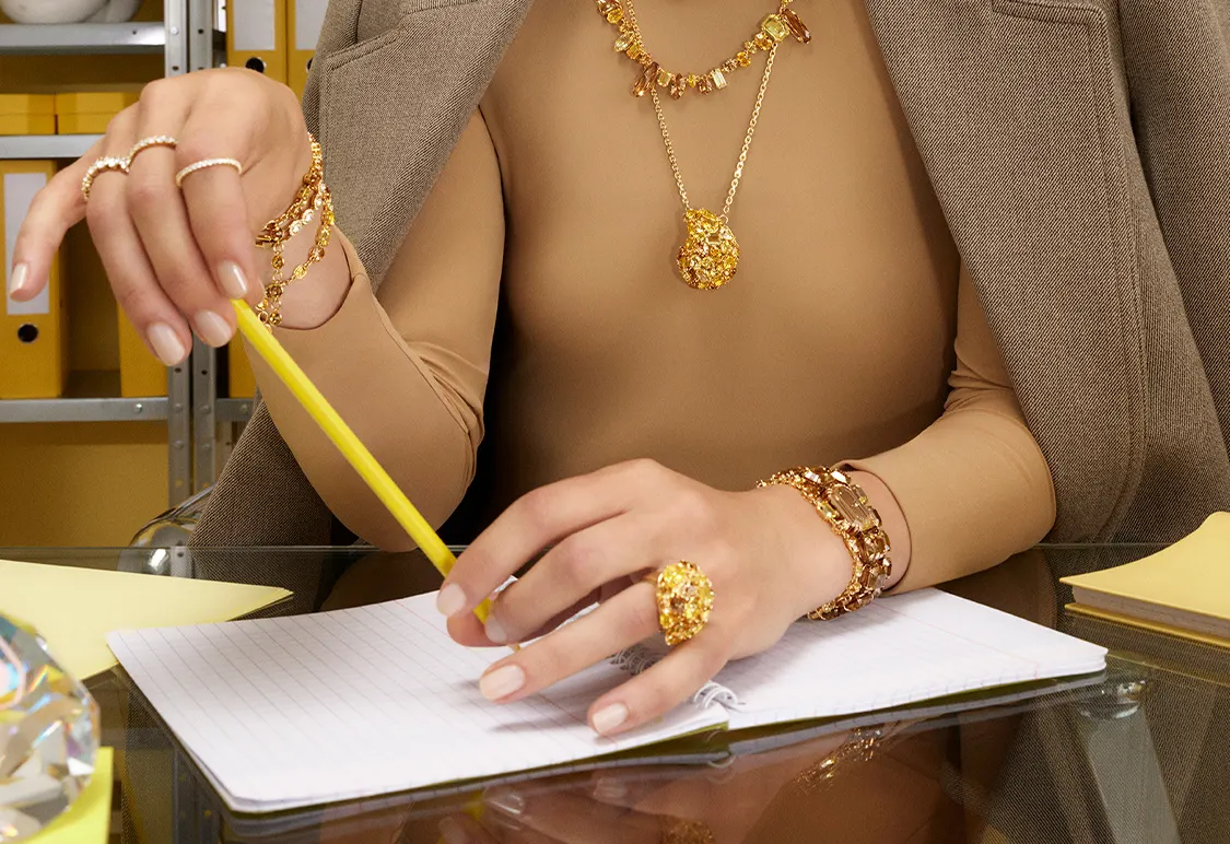 A model wearing gold tone Swarovski jewelry sits at a desk with a pad of paper and holds a yellow pen