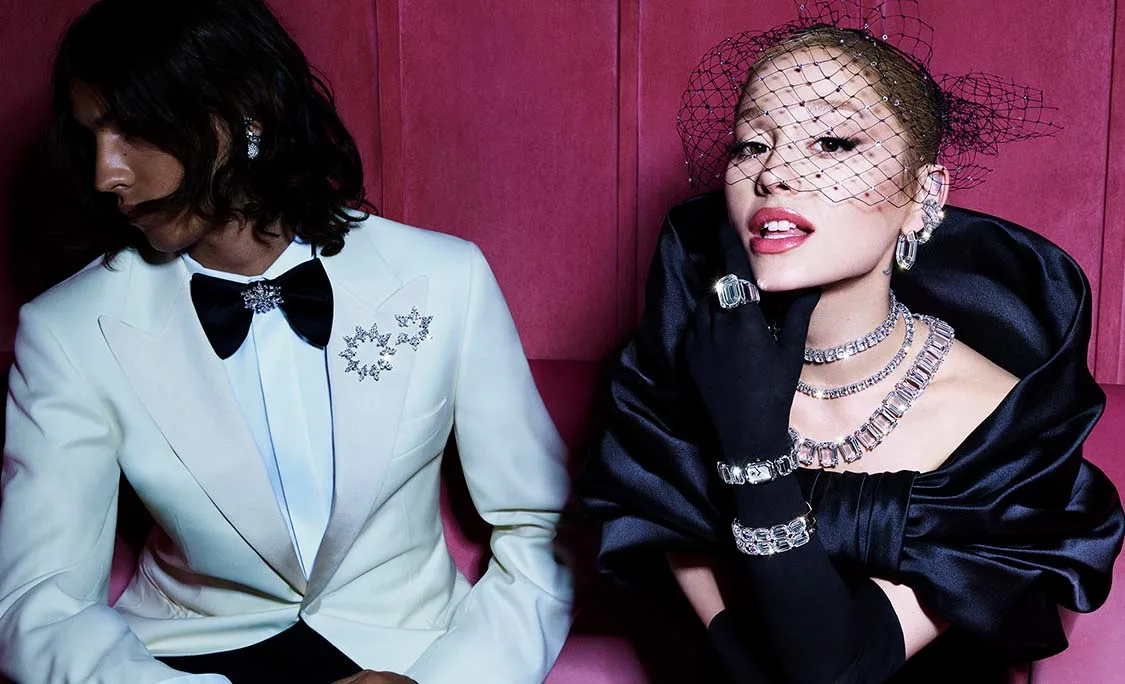 Ariana Grande in black outfit and Swarovski jewelry sitting next to man in white tuxedo