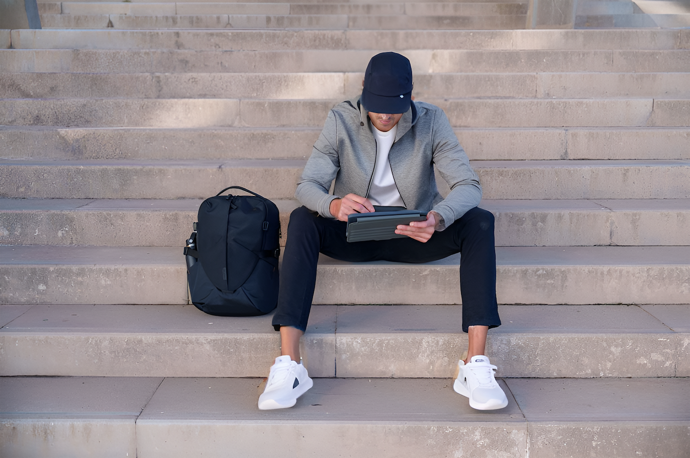 Modern man sitting on steps with his Targus black terra backpack looking at this tablet wrapped in a Targus tablet case. 