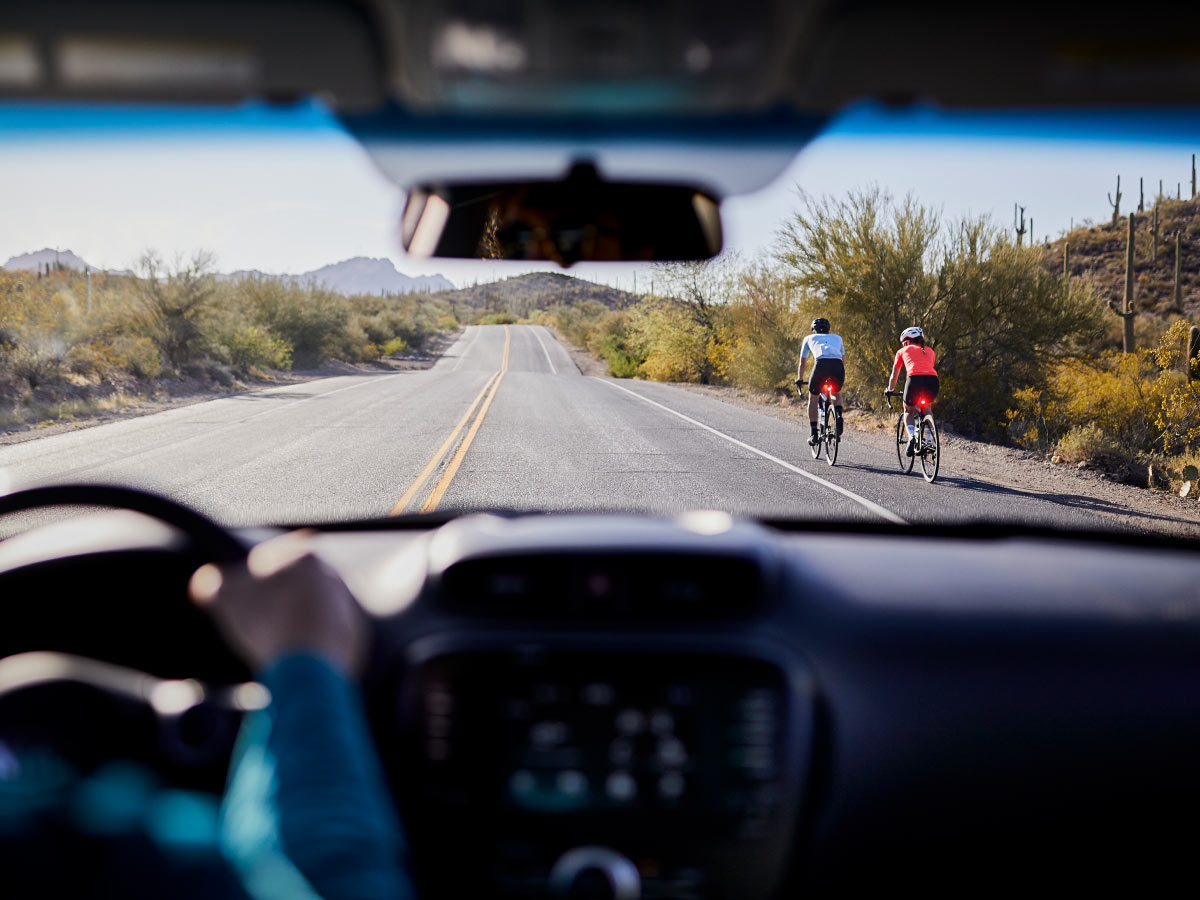 Person driving car on paved road and cyclists in front wearing helmets while riding bikes with daytime running lights turned on