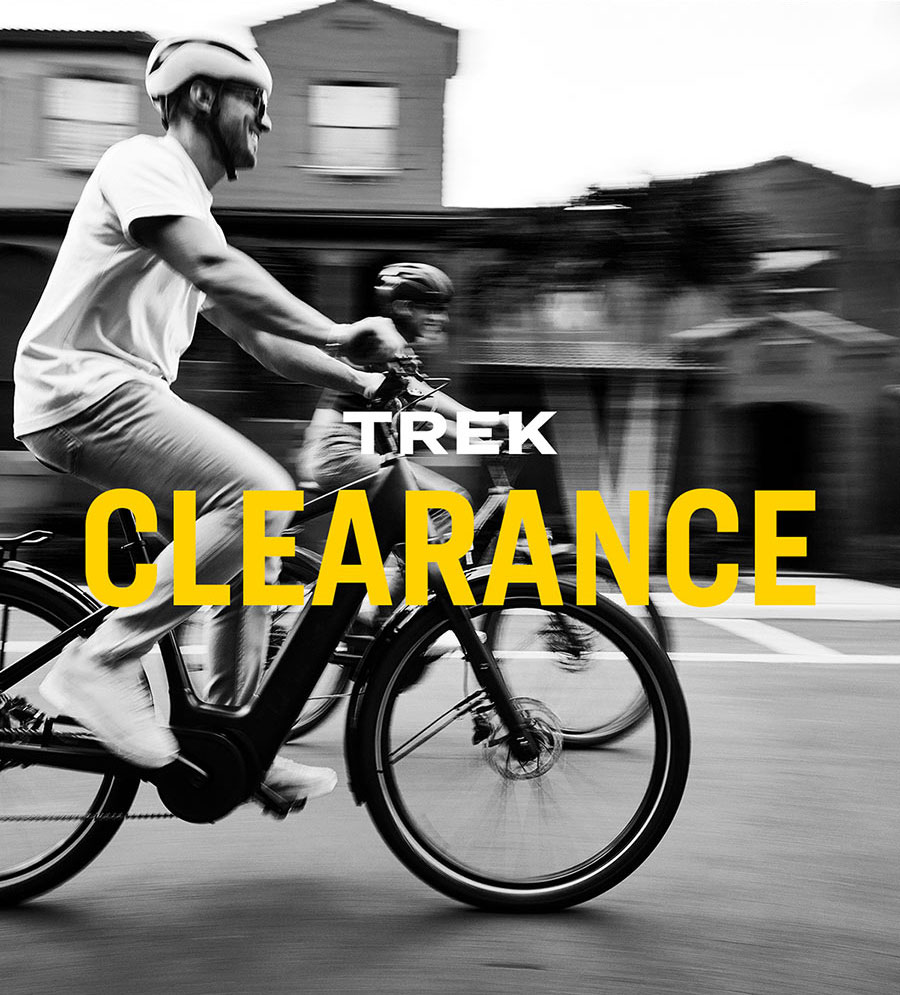 The words, 'TREK CLEARANCE' overlaying black and white photo of cyclists wearing helmets while riding bikes on paved road with houses in background