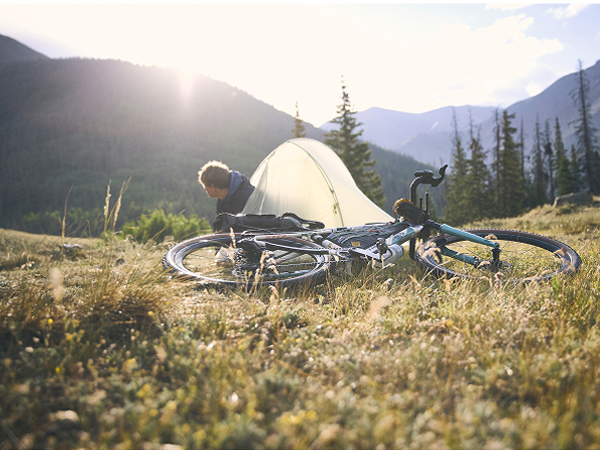 Cyclist peaking out of tent with all-new CheckOUT bike laying next to it and sun shining over mountains and trees
